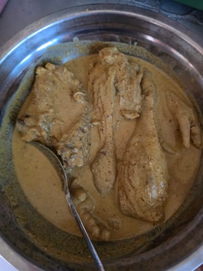 A close-up, overhead shot shows a large, shallow metal bowl filled with a creamy, light brown stew. Several pieces of chicken are submerged in the stew, including two legs and what appears to be a portion of a breast. A silver spoon rests in the bowl, partially submerged in the liquid, with its handle extending out of the frame. The stew has a textured appearance with small dark specks throughout. The bowl is rimmed and sits on a surface that is out of focus, but appears to be a countertop or table. The scene is likely indoors, given the lighting and the presence of kitchenware. The image was taken in Pintomakmur, Indonesia.