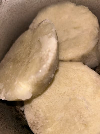 Three slices of cooked yam dominate the frame. They are pale, almost white, with a slightly grainy texture. The slices are irregularly shaped and stacked together. The background is a dark, undefined space, possibly a bowl or container holding the yam slices. Close proximity obscures any identifying context.