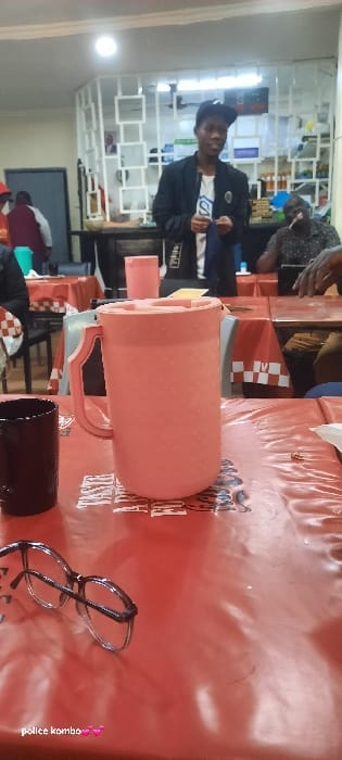 The scene depicts an indoor area of a restaurant or eatery located in Homa Bay, Kenya. In the foreground, a table covered with a red, somewhat wrinkled plastic tablecloth is visible. Centrally positioned on this table is a large, pale pink plastic water pitcher with a textured surface and a light-colored lid. To the left of the pitcher, a dark-colored cylindrical mug stands. A pair of black-framed eyeglasses rests on the lower-left portion of the tablecloth.
In the midground, a young male stands, facing slightly towards the right, with his hands engaged with a small, dark piece of fabric or paper. He wears a dark baseball cap positioned backward, a dark jacket over a white t-shirt featuring a dark "S" logo, and dark trousers. Behind him and to his right, a seated male with a patterned shirt is visible at a table covered by a red and white checkered tablecloth. This individual has his hands on the table, with a thin dark object near his mouth. Additional, partially obscured individuals are visible at tables to the left and further in the background.
The background elements include a white, open-shelf partition or room divider. Behind this partition, a counter area is discernible, displaying bottles and other items. The space is illuminated by overhead lighting, including a prominent circular ceiling light. The environment indicates an active, informal dining or social establishment.