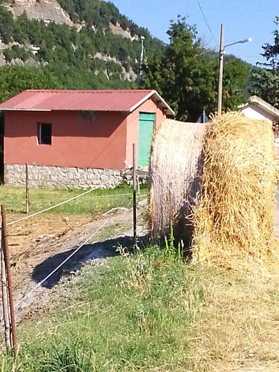 The image depicts an outdoor scene in Valle Castellana, Italy, featuring a rustic setting. In the foreground, a large bale of hay, golden brown and textured, rests on the ground. A wire fence with metal posts runs horizontally across the frame, separating the hay bale from a patch of rough terrain. Beyond the fence, a small, single-story building with a red metal roof and pale orange walls is visible. It has a small, dark window and a green door. The base of the building is constructed with grey stones. Behind the building, a lush green forest covers a steep, rocky hillside. In the background, a utility pole with a streetlamp stands tall against a clear blue sky. The overall impression is one of a rural Italian landscape on a sunny day.