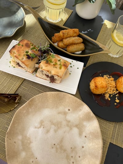 A close-up, overhead shot captures a table setting with various food items and drinks. In the foreground, a beige, irregularly shaped plate is empty, positioned on a woven, gold-colored placemat. To the left of this plate, a pair of tortoiseshell sunglasses rests on the placemat.

Behind the sunglasses and plate, a white rectangular plate holds a halved sandwich on focaccia bread, garnished with chives and microgreens. The sandwich filling appears to be shredded meat. Adjacent to this, a dark, leaf-shaped dish contains several golden-brown, fried croquettes served on a white sauce.

Further to the right, a black circular plate presents two golden-brown, round fritters or arancini, accompanied by a dark sauce and a sprinkle of crumbled breadcrumbs. Two glasses of lemonade, each with a lemon slice, are visible, one behind the croquettes and another partially visible at the top right. The setting is outdoors, suggested by the dappled sunlight and the greenery of a plant in a white pot in the background. This scene is likely set at a restaurant in the charming town of Pollença, Spain, known for its picturesque streets and culinary offerings.