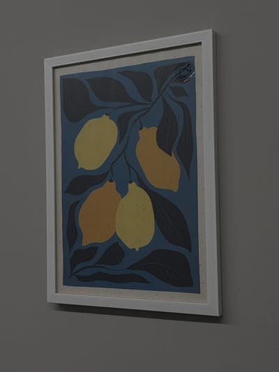 A framed print hangs on a wall. The print features four stylized lemons in various shades of yellow and orange, surrounded by dark blue-grey leaves against a blue background. The frame is white. The wall is painted a neutral color. The location in Zagarancea, Moldova is unknown.