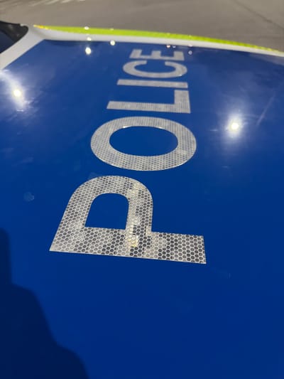 The image shows the word "POLICE" written in large, reflective letters on the blue surface of what appears to be a vehicle. The letters are silver and have a hexagonal pattern, indicating they are made of reflective material. The word is oriented diagonally from the top left to the bottom right of the frame. The background is a blurry, neutral color, possibly asphalt or a road surface. There is a green and yellow stripe along the top edge of the blue surface, likely part of the vehicle's livery. A shadow of a person is visible in the bottom left corner of the image, suggesting the photo was taken by someone. The scene does not provide any specific details about the city of Veliki Preslav, Bulgaria, as it focuses solely on the text on the vehicle.
