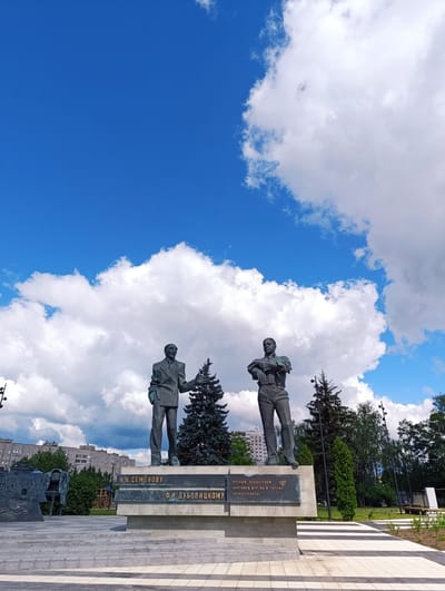 This media file captures a monument dedicated to N.N. Semenov and F.I. Dubovitsky, situated in Chernogolovka, Russia. The monument features two bronze statues of men standing side-by-side on a concrete pedestal. The man on the left is dressed in a suit and tie, while the man on the right wears a more casual outfit with a shirt and trousers. A dark green fir tree stands between them. The pedestal bears inscriptions in Russian, including "Н.Н. СЕМЁНОВУ" and "Ф.И. ДУБОВИЦКОМУ," and also "УЧЁНЫМ, СОЗДАТЕЛЯМ НАУЧНОГО ЦЕНТРА И ГОРОДА ЧЕРНОГОЛОВКА".

The monument is set against a backdrop of a bright blue sky filled with fluffy white clouds, suggesting fair weather and a daytime setting. In the background, there are tall trees and a multi-story residential building, indicating an urban or semi-urban environment. The ground around the monument is paved with light and dark striped tiles. The overall scene conveys a sense of civic pride and remembrance, honoring influential scientists and creators of the scientific center and the town.