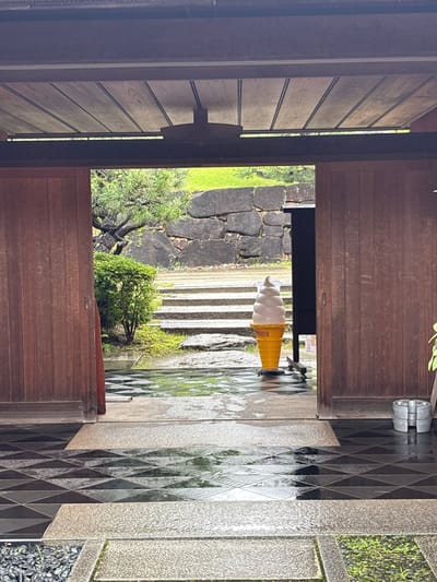 The image depicts an entranceway to a garden area, likely in Nagakute, Japan, given the context provided. The scene is observed from an interior space looking out through an open wooden doorway. A large, orange ice cream cone statue, presumably a soft-serve vanilla, stands prominently in the middle ground on a wheeled base. Behind the statue, a pathway with stone steps leads up towards a rough-hewn stone wall. Lush green bushes and a mature pine tree are visible to the left of the steps. The ground in front of the doorway is paved with a checkered pattern of black and light-colored tiles, which appear wet, suggesting recent rain. The overall atmosphere is serene and hints at a Japanese garden setting, possibly at a cafe or tourist attraction. There are no people visible in the image.