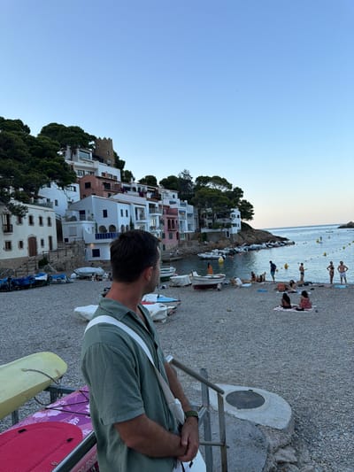 The image depicts a beach in the town of Begur, Spain, situated on the Costa Brava. The foreground shows a man in a green shirt looking out towards the sea. To his left, there are two paddleboards on a rack. The beach itself is pebbled and lined with several small boats pulled ashore. In the background, a cluster of white and pastel-colored buildings cascades down a hillside, topped with lush green trees. A small, stone tower, a remnant of medieval fortifications, is visible among the buildings. The sea is calm and blue, with a few people swimming or sunbathing on the beach and a paddleboarder in the water. The sky is clear and light blue, suggesting it is either late afternoon or early morning. The overall scene conveys a peaceful, seaside holiday atmosphere.
