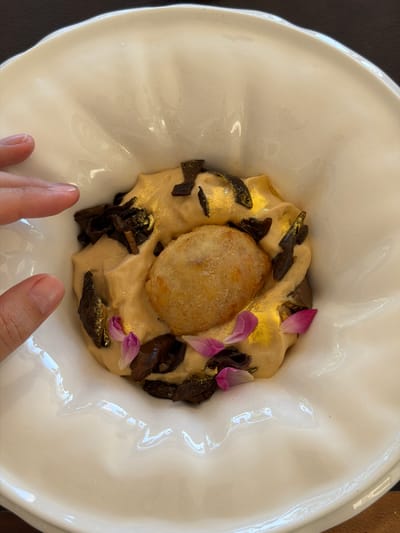 In Baan Thong Krut, Thailand, a close-up, overhead shot captures a white, scalloped bowl of food. A person's left hand, with manicured nails, rests on the rim of the bowl. The dish itself features a creamy, golden-hued base topped with dark, irregular pieces that appear to be food items, possibly truffles or mushrooms, and delicate pink petals. A golden dusting is visible on the creamy base and some of the darker toppings. In the center of the dish is a single, golden-brown, fried or battered item, possibly a croquette or a small pastry. The bowl rests on a dark surface, likely a table.