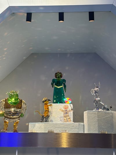 The image shows a display case with four crystal figurines arranged on stepped platforms. The figurines, from top to bottom, are: a dog, a figure in green with a lollipop, a figure in gold, and another figure in green. The stepped platforms on which the figures are placed are marked with numbers that appear to be product or model numbers. The scene is contained within a glass display case located in Spreitenbach, Switzerland.