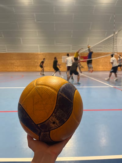 The image shows a person holding a yellow and dark blue handball in their right hand. In the background, a handball training session is in progress on an indoor court. Several players are visible in motion, jumping and reaching for the ball near a handball net. The court has a blue surface with red and white lines. The environment appears to be a gymnasium or sports hall. The location is most likely near Pobrdje, Montenegro. The scene suggests a typical handball training session in a sports facility.