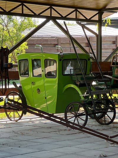 A bright green horse-drawn carriage is parked under a wooden and metal roof structure. The carriage has four windows with draped curtains and two lanterns attached to the exterior. It is supported by two large spoked wheels at the rear and two smaller spoked wheels at the front. In front of the carriage, a metal frame with a seat and more spoked wheels suggests another, smaller carriage or vehicle. The ground is paved with square tiles. In the background, a wooden fence and a glimpse of trees and a building can be seen. The scene appears to be outdoors, possibly in a rural or historical setting. There are no people present in the image.