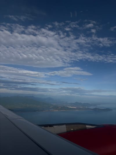 The image displays an aerial view from an airplane window, looking out over a coastal landscape under a bright, partly cloudy sky. In the lower right foreground, a portion of the airplane's red engine nacelle is visible, alongside the grey and white leading edge of the wing. The main body of the wing appears dark grey.
The sky occupies a significant part of the frame, showing a clear blue expanse dotted with scattered white clouds, ranging from small wisps to larger, more defined formations. The bright illumination indicates daytime conditions.
Beyond the aircraft, a vast landscape unfolds. A large body of water, likely a sea or a significant lagoon, stretches across the mid-ground. It is bordered by a long, narrow strip of land that separates it from what appears to be another body of water or the open sea further out. Hazy green mountains rise from the land, diminishing in clarity towards the horizon. Along the coastline, some light-colored patches suggest developed or urbanized areas. Given the provided context, the scene likely depicts the coastal area of Huế, Vietnam, with its characteristic intermingling of water, land, and mountains. No visible text is present in the image.