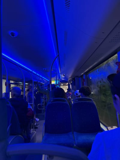 The image depicts the interior of a bus, illuminated by blue LED lighting. Several passengers are seated, facing forward. A security camera is visible on the ceiling. The bus is likely in motion, as indicated by the blurred exterior view through the windows. The setting is described as being in Campitello di Fassa, Italy, though this information is external to the visual content of the image.