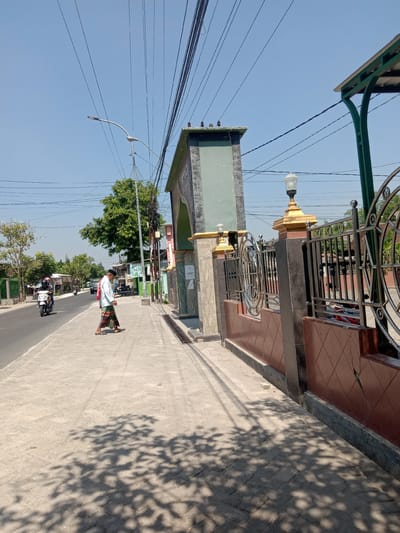 The image depicts a sunny street scene in Balongrejo, Indonesia. In the foreground, a man wearing a white shirt and a sarong walks on a paved sidewalk. To the right of the sidewalk is a brown tiled wall topped with a decorative metal fence. A large, arched gateway with a green facade stands further back, with a street lamp on a golden base positioned in front of it. Overhead, a tangle of power lines crisscrosses the bright blue sky. A road runs parallel to the sidewalk, with a motorcycle and a car visible in the distance. Trees and buildings line the street. The overall impression is that of a typical day in a residential or commercial area.