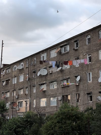 The image depicts a building with multiple stories, likely an apartment complex, in Shaki, Azerbaijan. Laundry is hanging on clotheslines strung between balconies or windows, suggesting domestic activity. Various satellite dishes are mounted on the exterior walls, indicating modern amenities. The building's facade appears weathered, typical of older construction. Green trees and foliage are visible on the right side of the frame, partially obscuring some of the building and suggesting an outdoor setting. The sky is overcast, hinting at cloudy or perhaps rainy weather. A bird is in flight in the sky on the left side of the image. The overall scene is mundane, showcasing everyday life in an urban or semi-urban environment. There is no visible text in the image.