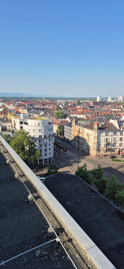 An aerial view captures a European city, likely Karlsruhe, Germany, on a clear, sunny day. The foreground shows the flat, dark rooftop of a building, equipped with a metal railing and wire safety system, anchored by several rounded, dark objects. Beyond the rooftop, the scene unfolds into a dense urban landscape characterized by a mix of building architectures.
Dominating the mid-ground is a striking, curved white building with multiple levels of windows, featuring a green roof. Adjacent to it, older, multi-story buildings with terracotta roofs and cream-colored facades line the streets. A yellow and black tram is visible on the street, suggesting public transportation is in use. The street layout indicates intersections with tram tracks.
The background reveals a sprawling city with numerous buildings featuring red-tiled roofs, creating a homogeneous urban texture. In the far distance, a range of low mountains or hills can be seen under a clear blue sky, with some taller, modern buildings interspersed within the city skyline. The overall impression is one of a well-established European city with a blend of historical and modern architecture, bathed in the bright light of a summer day.