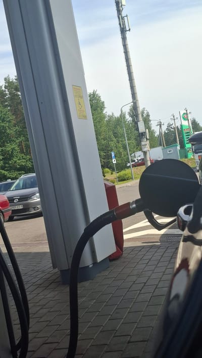 An outdoor shot captures a gas pump nozzle connected to a car's fuel filler on a sunny day. The scene is set at a gas station, possibly in the Драчкаўскі сельскі Савет, Belarus, as indicated by the Cyrillic text on a yellow sticker on the gas pump which reads "Внимание" (Attention). The sticker also contains a phone number: "+375 17 2093539" and a wheelchair symbol, suggesting accessibility information.
In the background, trees and a cell tower are visible. A dark-colored Volkswagen station wagon is parked nearby, displaying a license plate "0906 AB-8". The ground is paved with interlocking gray bricks. The composition is framed by the gas pump column on the left and the car's side mirror and fuel cap area on the right. The overall lighting suggests daytime with clear skies.