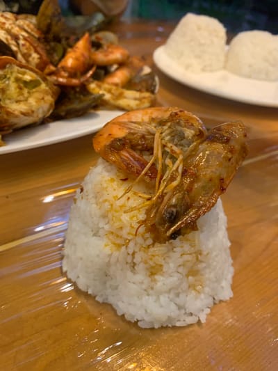 A close-up, overhead shot captures a seafood dish served on a wooden table, partially covered with clear plastic wrap. The main subject is a large, cooked shrimp seasoned with spices, placed atop a mound of white rice. To the left, another plate holds a collection of seafood, including what appear to be crab legs and more shrimp, all coated in a similar reddish-brown sauce. In the upper left corner, two servings of plain white rice are visible on a white plate. The lighting suggests an indoor setting, possibly a restaurant, with a warm ambiance. The image does not contain any visible text. The overall impression is a hearty, flavorful meal.