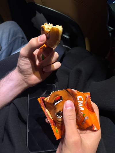 The image depicts a person indoors, likely in a car, holding an opened orange snack package in their right hand and a partially eaten snack in their left. The snack package is branded "Grand Casteli CMAK." A black smartphone or tablet lies flat beneath the snack package. The person is wearing blue jeans and a black top. The background includes a glimpse of a car interior. There is no information in the image to identify the location as Tsakhkadzor, Armenia.