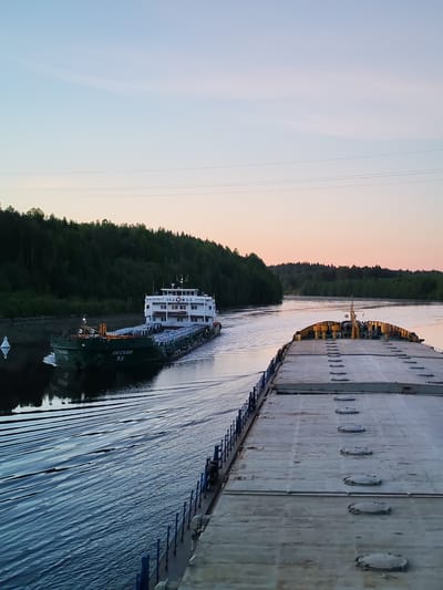 The image captures a peaceful scene on a waterway, likely a river or canal, at either dawn or dusk. The sky displays a gentle gradient from pale blue at the top to soft pink and orange closer to the horizon, indicating early morning or late evening light.
In the mid-ground, a dark green cargo vessel with a prominent white superstructure is navigating the calm waters, creating a visible wake. The name "ОКСКИЙ" (Okskiy) and the number "53" are clearly inscribed on its hull in white. The superstructure also bears the text "ОКСКИЙ И-53". This vessel appears to be a river barge. A white conical buoy is visible to the left of the passing ship.
The photograph is taken from the perspective of another large vessel, the deck of which dominates the foreground on the right side of the image. This deck is flat, grey, and features a series of circular, sealed hatches, bordered by a blue railing. This vessel also appears to be a cargo barge.
Both sides of the waterway are lined with dense, dark green forest, suggesting a natural or canalized environment cutting through a forested area. Overhead power lines are faintly visible against the sky. The water is smooth, reflecting the surrounding landscape and sky. The visible Russian text on the ship, "ОКСКИЙ", points to a location within Russia, such as Depo. The scene evokes a sense of tranquil passage through a serene natural setting.
