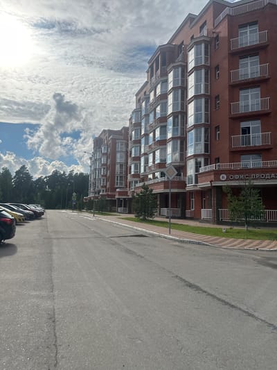 The image shows a row of brick apartment buildings with many windows and balconies. In front of the buildings is a paved road with parked cars on the left side. A sidewalk with grass and trees borders the road in front of the buildings. A sign on the building says "ОФИС ПРОДАЖ," which translates to "Sales Office." The sky is cloudy with patches of blue. The location appears to be in Snt Del'fin, Russia, based on the text.