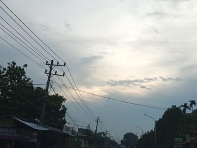 The image depicts a street scene in Gampongrayadagang, Indonesia, under an overcast sky. Power lines are a prominent feature, strung between multiple utility poles that line the street. Some of these poles are equipped with insulators to hold the wires. On the left side of the image, a low-slung building with a tin roof is visible, appearing to be a storefront or stall. Text on the building, though partially obscured and blurred, includes the words "NGAN KUAT" and "ASI KEMBALI LANCAR". Lush green trees, including some palm trees, are scattered throughout the scene, creating a natural contrast with the man-made infrastructure. Several streetlights are also present, suggesting the area is illuminated at night. The overall impression is one of a typical semi-urban or rural street in Indonesia, characterized by its electrical infrastructure and abundant vegetation. There are no people or vehicles clearly visible in the frame.