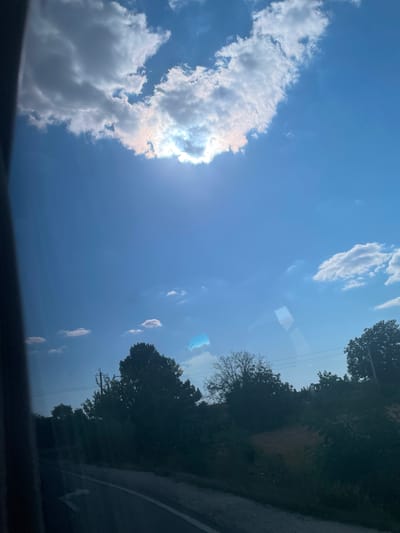 The image depicts a view of the sky and the landscape from inside a vehicle, likely on a road. The sky is a vibrant blue with scattered clouds, some of which are illuminated by the sun, creating a bright halo effect. The sun is partially obscured by a large, fluffy cloud, casting a warm glow. Below the sky, a treeline is visible, with various types of trees, some dense and leafy, others bare. A road with a white arrow marking can be seen in the foreground, suggesting a journey or travel. The overall impression is of a sunny day in a rural or suburban setting.