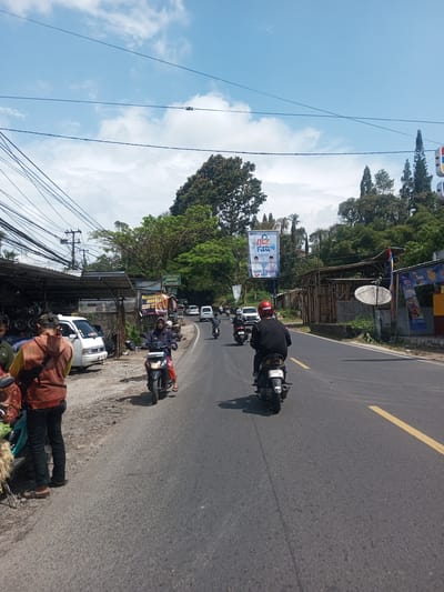 The image depicts a street scene in a location with tropical vegetation and overhead power lines. Several people are present; some are riding motorcycles on a paved road, while others are standing beside the road, possibly near shops or vendors. The sky is blue with some clouds. A political billboard is visible on the side of the road. The location is not Zeyathiri District, Myanmar. The license plate on the motorcycle is not from Myanmar.
