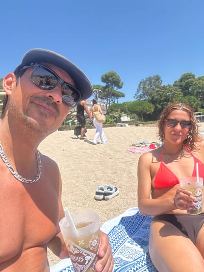 In the city of Castell d'Aro, Platja d'Aro i s'Agaró, Spain, two individuals are captured in a selfie on a beach under a clear blue sky. A man wearing a dark flat cap and sunglasses is positioned in the foreground on the left, holding a drink with a straw. He is topless and adorned with a thick silver chain necklace. To his right, a woman in a red bikini top and dark bottoms is also holding a drink with a straw, her hair appearing wet. She is wearing sunglasses and a gold chain necklace. The background reveals a sandy beach with scattered beachgoers, trees, and a body of water. A patterned beach towel is visible beneath the couple. The overall scene depicts a casual, sunny day at the beach.