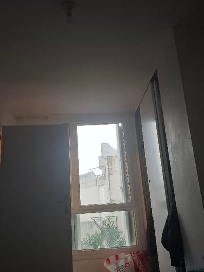 The image presents an interior view of a room in Saint-Martin-d'Hères, France, looking outwards through a rectangular window. The room itself is dimly lit, featuring plain, light-colored walls and a ceiling. A small, indistinct light fixture or switch is visible on the left wall. Above the window, a structural beam or track is present, from which a dark piece of clothing or fabric hangs.
The window has a white frame, divided into several panes, and provides a view of the exterior. The outdoor scene is brightly illuminated, indicating it is daytime, likely with overcast weather as the light appears diffused and details are somewhat washed out. Through the window, parts of light-colored, multi-story buildings, typical of an urban or suburban setting, are visible. Some green foliage, possibly from a tree, can be seen to the right through the window.
Partially visible on the right side of the window is a white and red plastic shopping bag. Text on the bag appears to read "MARCHÉ", or possibly "MARCHÉ PLUS", suggesting it is a shopping bag. Below the window, a large, dark, flat surface occupies the lower portion of the image. No people or specific activities are depicted, giving the scene a quiet, static quality. The strong contrast between the dim interior and the bright exterior through the window is a notable lighting detail.