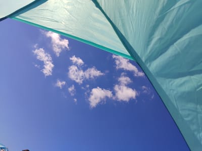 The image captures a low-angle view of a bright blue sky dotted with fluffy white clouds, framed by the turquoise fabric of what appears to be a tent or awning. The fabric, with its visible creases and seams, occupies the upper right portion of the frame, extending diagonally across. The sky itself is clear and expansive, with the clouds appearing scattered and thin, suggesting a fair-weather day. While the image is devoid of any identifiable landmarks or architectural features that would definitively place it in Kandıra, Turkey, the overall scene evokes a sense of outdoor leisure or camping in a location that experiences such clear skies and cloud formations. There are no people or specific actions depicted in the image.