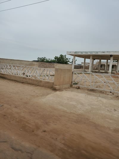 The image shows an unfinished concrete structure with pillars and a roof, possibly a gas station or a covered waiting area. There is a white metal fence in the foreground and a dirt road in the lower portion of the image. In the background, there are more unfinished buildings made of concrete blocks and some trees. The sky is overcast. The text visible on the roof appears to read "IMPORT OIL AND GAS LTD". The location is identified as Kwadon, Nigeria. There are no people visible in the image.