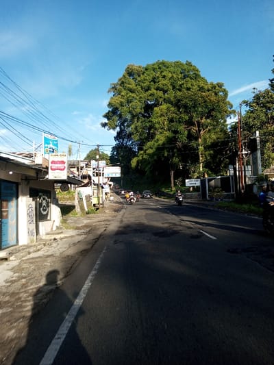 A daylight street scene depicts a paved road with an adjacent concrete sidewalk under a clear blue sky. On the left, a row of low-rise commercial buildings features a light-colored facade with graffiti "RWHS" and multiple overhead utility lines. A prominent sign for a dental clinic displays red lips and white teeth, with the text "BUMI GIGI." The asphalt road, showing dark patches and a faded white centerline, extends into the distance, gently curving to the right.
Multiple motorcycles and at least one car are observed traveling away from the viewer on the road. A white directional sign on the right side of the road points left, bearing the text "JALAN ST. YUSUF." Lush green trees form a dense canopy along the right side of the street and in the background. Long shadows, including that of the photographer on the lower-left sidewalk, are cast by the sun. Based on the visible text "BUMI GIGI" and "JALAN ST. YUSUF," the scene is located in an Indonesian or Malay-speaking region, not Khouathineung, Laos.