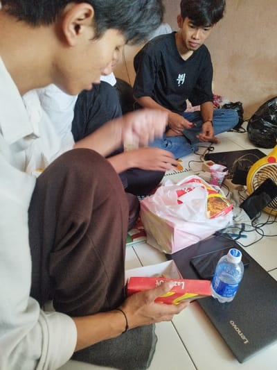 In an indoor setting in Muka, Indonesia, three males are seated on a light-colored tiled floor with a plain, light-brown wall in the background.
In the left foreground, a male with dark hair and a light-colored collared shirt is partially visible, facing right. He wears dark pants and holds a red "Lazat" branded rectangular food box in his hands. His right arm is visible, showing a black string bracelet on his wrist, and his left hand is blurred due to motion.
In the middle background, a male with short dark hair and a light beard sits cross-legged, wearing a black t-shirt with a white character design and ripped blue jeans. He looks towards the left, holding a small red object in his hands, appearing to manipulate it.
A third, less distinct individual is partially visible behind the foreground male.
Various items are arranged on the floor between the individuals:
* A grey Lenovo laptop is positioned on the right.
* A clear plastic bottle of mineral water with a blue cap rests on the laptop.
* A large white plastic bag contains a pink box and other items, including a package of instant noodles.
* To the right of the plastic bag, a white power strip with multiple plugs is visible amidst tangled black cables.
* A small yellow desk fan is situated near the power strip.
* Several small plastic cups and containers are scattered nearby.
* A crumpled black plastic bag is visible in the far right background.
The scene depicts a casual gathering with food and electronics spread across the floor.