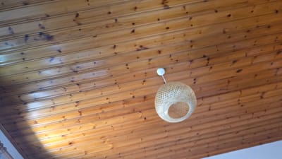 The media file displays a close-up, low-angle shot of a wooden paneled ceiling with a woven light fixture. The ceiling is made of horizontal wooden planks, stained a warm, honey-brown color and featuring natural knots and grain patterns. A prominent shadow, possibly cast by a window or skylight, falls diagonally across the upper right portion of the image, creating bright highlights on the wood.
Hanging from the ceiling is a light fixture crafted from woven natural material, possibly rattan or bamboo. It has a spherical, open-weave design with a circular opening at the top. A thin white cord extends from the bottom of the light fixture to a small, white circular object, likely a switch or ballast. The light fixture is positioned off-center in the frame, towards the left.
The scene is static, with no activity taking place. The lighting suggests it could be daytime, given the strong shadow and highlights, though the exact time of day is not definitively ascertainable. The setting appears to be indoors, likely a room with a distinctive wooden ceiling. No people are visible, and no text is present in the image. The overall impression is one of simple, natural interior design.