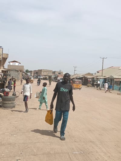 This image depicts a bustling street scene in Hotoro South, Nigeria, under a bright, hazy sky. The foreground features a dark-skinned adult male walking towards the left side of the frame, holding a yellow plastic bag in his left hand. He wears a dark grey t-shirt with a light green "Ziggo" logo, blue trousers, and grey sandals, and is looking directly at the viewer. To his left, in the midground, a young male in a white long-sleeved top and dark shorts walks left, past a stack of four black tires and a red-and-silver generator unit. Another young male in a light green traditional outfit, carrying a white bucket, walks further left, behind the first young male.
The unpaved dirt road shows visible tire tracks. In the mid-background, a yellow auto-rickshaw (Keke Napep) travels away from the viewer on the right side of the road, while a person on a motorcycle and another on a bicycle are visible further down the street on the left. Buildings line both sides of the road, ranging from single-story structures with corrugated metal roofs to multi-story buildings, suggesting a mix of residential and commercial use. Several utility poles with overhead wires are present, particularly on the right side of the road, which appears to be lined with market stalls or shops. Numerous other pedestrians are visible in the background, including individuals in traditional white garments on the right. Shadows cast by the subjects suggest a high sun position.