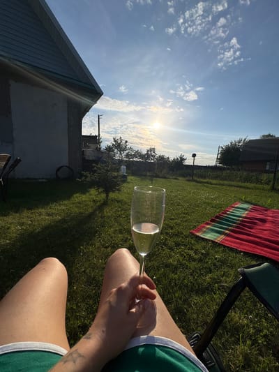 A first-person perspective shot shows a person relaxing in a chair outdoors in what appears to be a rural setting near the city of Nemantsy, Belarus. The sun is setting or rising, casting long shadows across a grassy lawn. In the foreground, the person's legs are visible, clad in green shorts, and a hand holds a stemmed glass filled with a light-colored liquid, likely champagne or white wine. To the right, a red and green striped blanket is spread on the grass. In the background, a small evergreen tree stands near a rustic-looking building with a grey facade and a dark, sloped roof. Further back, a fence encloses a garden area, and more buildings with dark roofs are visible, characteristic of a rural Belarusian village. The sky is blue with scattered white clouds, and the sun is a bright orb near the horizon, creating a warm, golden light.