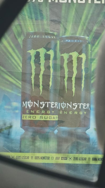 The image shows two stacked cans of Monster Energy Zero Sugar drink, viewed through a speckled surface. The cans are primarily black with the iconic green Monster Energy claw logo prominently displayed on each. Vertical text on the front of the cans reads "MONSTERIONSTER ENERGY" and "ZERO SUGAR". One can also has the word "TAURIN" visible vertically. A yellow stripe runs vertically along the right side of the image with text that includes "ZERO AZÚCAR 100% MONSTER". The background behind the cans appears to be a green, abstract design with radiating lines. The overall impression is of product advertising, likely in a retail or display setting. There are no people or specific location cues beyond the Spanish text on the yellow stripe. The lighting suggests an indoor environment.