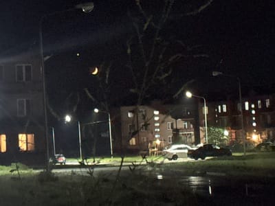 The image depicts a nighttime scene in the city of Malgobek, Russia. In the sky, a crescent moon and a bright celestial body, likely a planet or star, are visible. In the foreground, a dark, grassy area with some foliage is present, with a wet surface indicating recent rain. Beyond this, a road is visible, with several cars parked or driving. On the left side of the road, a multi-story building with illuminated windows is present. Further down the road, more multi-story buildings with lit windows are visible. Streetlights illuminate parts of the scene.