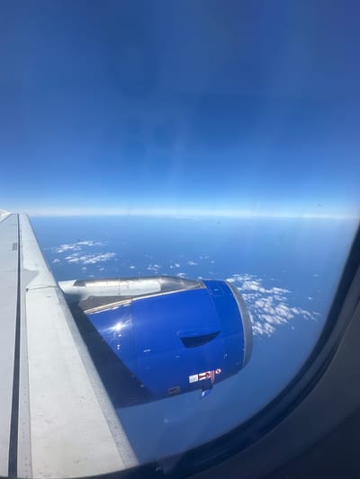 An aerial view from an airplane window shows a bright blue sky over a vast expanse of ocean. White, puffy clouds are scattered across the water's surface. A portion of the airplane's wing, made of light gray metal, is visible on the left side of the frame. To the right of the wing, a blue-colored jet engine is prominent, with a metallic section just behind it. The sun is shining brightly, creating a reflection on the engine. The image does not depict the city of Viehhausen, Austria; it shows an oceanic landscape from a high altitude.