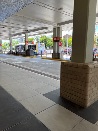 This image captures a modern, covered bus interchange or transport hub, likely in Indooroopilly, Australia, under bright daytime conditions. The perspective is from within the sheltered passenger area, looking out towards the bus lanes and the street beyond.

The foreground features a sturdy, light-brown brick pillar on the right and several smooth, grey-beige pillars supporting the white overhead roof structure. The ground is paved with large square tiles in alternating light grey and dark grey shades, forming a diagonal pattern. A long strip of tactile paving or a metallic grate runs along the edge of the pedestrian area, separating it from the bus lane.

Beyond the shelter, a designated bus lane is visible, marked with yellow lines and the word "STOP" painted in white on the asphalt. A low metal fence or barrier separates the waiting area from the road. Multiple tall signposts are present, each displaying various directional and informational signs. Notable among these are red octagonal "STOP" signs and rectangular signs featuring a red square with a white bus icon. One sign clearly shows an upward arrow, the letter "B", and smaller text "BUS ROUTE C". Another sign further down the lane indicates "A" with an upward arrow and a bus icon.

A bus, partially visible in the lane, features colorful advertising on its side, with the words "INNOVATION UNWRAPPED" discernible. Several people are seated on benches beneath the shelter, appearing to be passengers waiting for buses. The background reveals lush green trees and glimpses of distant buildings, suggesting an urban or suburban setting. The bright light indicates clear weather.