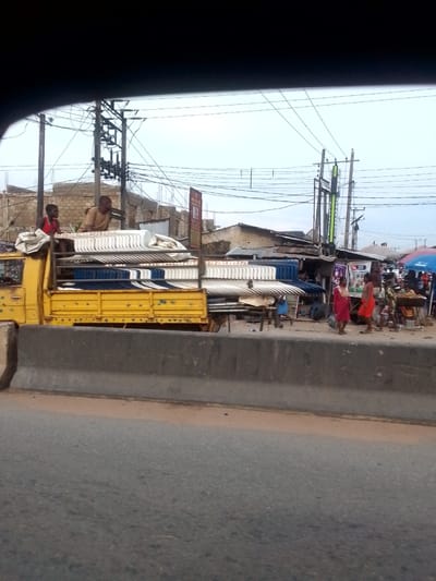 The image shows a street scene in Ihiala, Nigeria. A yellow truck overloaded with white and blue plastic chairs has two men sitting on top of the chairs. The truck is moving alongside a concrete divider. On the other side of the divider, people are walking and interacting with vendors at roadside stalls. Power lines crisscross overhead. The environment is urban with buildings and structures visible in the background.