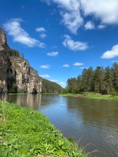 A landscape photograph taken in Kul'metovo, Russia. The image features a river running between a rocky cliff on the left and a grassy bank on the right. A line of trees sits atop the cliff. The sky above is blue with scattered white clouds. A red boat is visible in the distance on the river. The image is oriented with the top of the scene to the left.