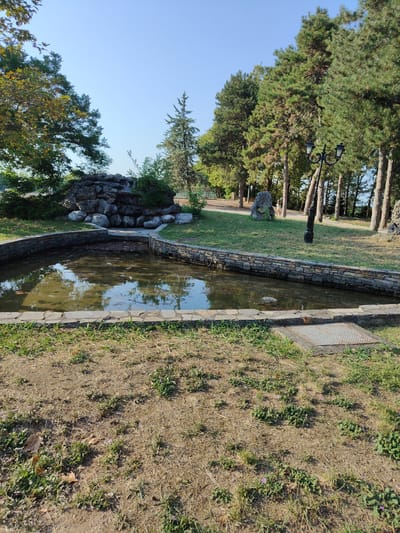The image shows a pond with a stone border, located in the city of Litochoro, Greece. Behind the pond, a rock formation with greenery simulates a waterfall. The area is surrounded by trees, including pine trees, and a clear blue sky is visible. A vintage-style street lamp stands near the trees. The ground in the foreground is covered with dry grass and some small plants. A metal grate is visible on the ground near the pond.
