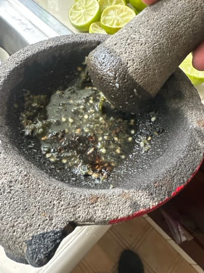 A stone mortar and pestle grind ingredients, likely for salsa. Seeds are visible within the mixture, submerged in a watery base. A hand holds the pestle above the mortar. Lime wedges sit nearby on a white surface. The mortar itself shows a worn texture. A red line is visible on the mortar. The scene is set on a countertop in what seems to be a kitchen setting.