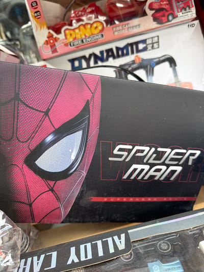 The image shows a close-up of various toy boxes, with a prominent Spiderman mask on a black box in the foreground. The Spiderman box is partially obscured by other items, including a clear plastic bag with some contents visible inside, and a black box with white lettering that reads "ALLOY CAR" in reverse. Above the Spiderman box, other toy boxes are visible, one with "DINO FIRE ENGINE" branding and another with "DYNAMIC" in large letters. The background appears to be a retail environment, possibly a market or shop, with shelves and various products. There is no indication of the city of Tukaytepa, Uzbekistan in the image.
