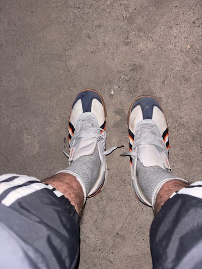 The image shows a top-down view of a person's legs and feet, wearing white athletic shoes with blue and orange accents, grey socks, and black athletic shorts with white stripes. The shoes appear to be on a concrete surface, which could be a sidewalk or pavement. The lighting suggests it might be daytime, though it's difficult to ascertain the exact time of day. There are no strong indicators of the weather. The setting is a generic outdoor environment, possibly urban. The person is standing still, with no specific activity or event clearly depicted. The focus is on the footwear and lower legs. The visible text on the shoes appears to be the brand name "PUMA". The location context of Saran city administration, Kazakhstan, is provided externally and is not visually evident in the image itself.