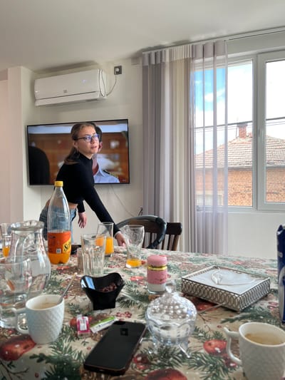 The image shows an indoor scene in Karavelovo, Bulgaria. A young woman with glasses is seated in front of a TV screen displaying a scene from a show. She is wearing a black shirt and jeans. On the table in front of her, there are several empty drinking glasses, a bottle of orange juice, a black smartphone, a box with a silver frame, and a mug. There is a window with sheer curtains in the background, and a white air conditioner mounted on the wall.