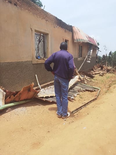 The image shows a man standing in front of a building with a partially collapsed roof in Kwal, Nigeria. The building is made of concrete and has a brown exterior. The roof is made of corrugated metal sheets, some of which have been blown off by the wind. The man is wearing blue jeans and a purple shirt. He is looking at the damaged roof. The ground is dirt and there are some pieces of broken metal lying around. The scene appears to have been impacted by a storm, likely with strong winds. The surrounding vegetation is sparse, suggesting a dry environment.