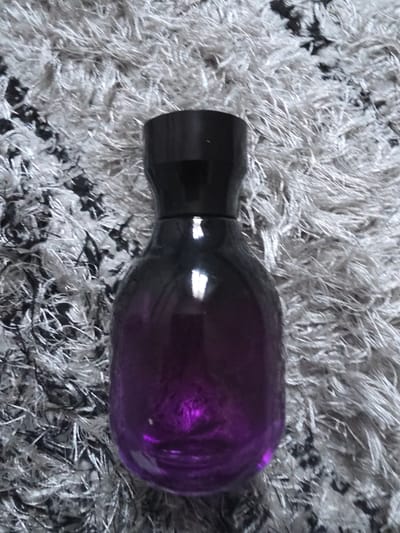 A purple, oblong-shaped glass bottle with a black cap lies horizontally on a gray and white shag rug. The bottle is oriented with its cap on the left and the wider body on the right. The surrounding environment consists solely of the rug's texture. No people, other objects, actions, or interactions are visible. The location is not identifiable from the image.
