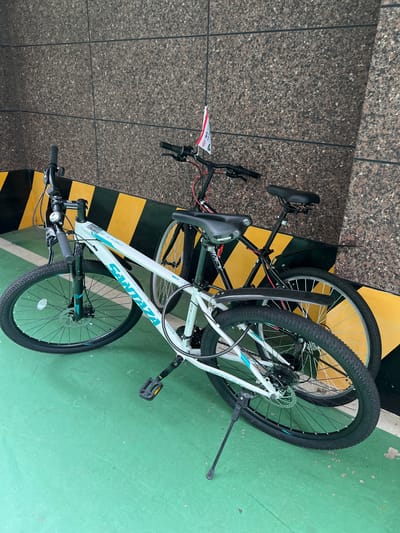 Two bicycles are parked next to each other in a covered area with a textured, speckled brown wall. The bicycle in the foreground is white with turquoise accents and the brand "SANTAZA" is visible on the frame. It has a kickstand and a disk brake on the front wheel. The second bicycle, behind the first, is black and has a small red and white flag attached to its handlebars. Both bicycles have knobby tires, suggesting they are mountain bikes. The ground is painted a bright green, and yellow and black hazard stripes are visible on a low wall or barrier behind the bicycles. The scene appears to be in an urban environment, likely a parking area or a covered walkway. The presence of the flag might indicate a local event or advertisement.